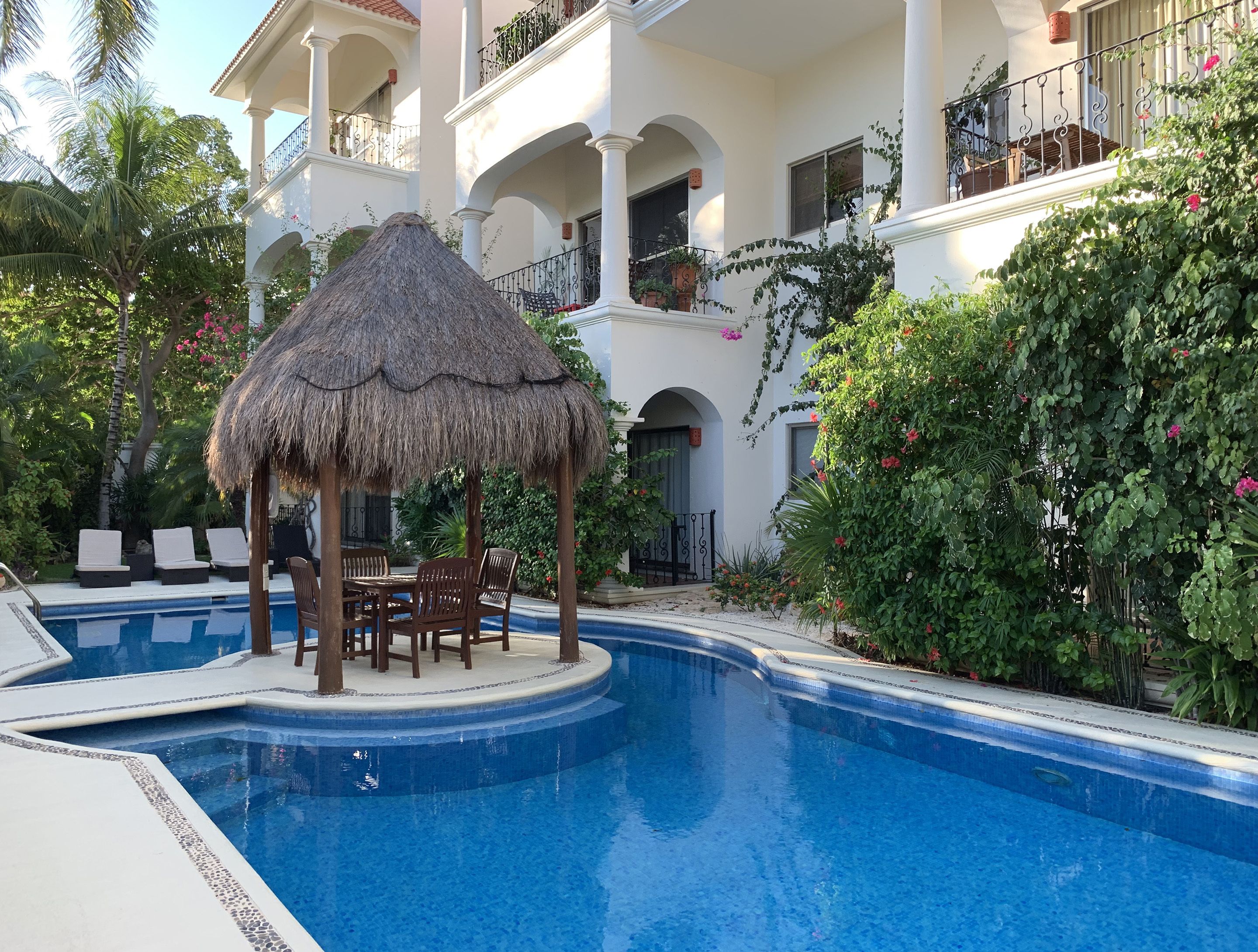 Ein Swimmingpool mit einem strohgedeckten Gazebo und Liegestühlen, umgeben von üppiger Vegetation und einem mehrstöckigen Gebäude mit Balkonen.