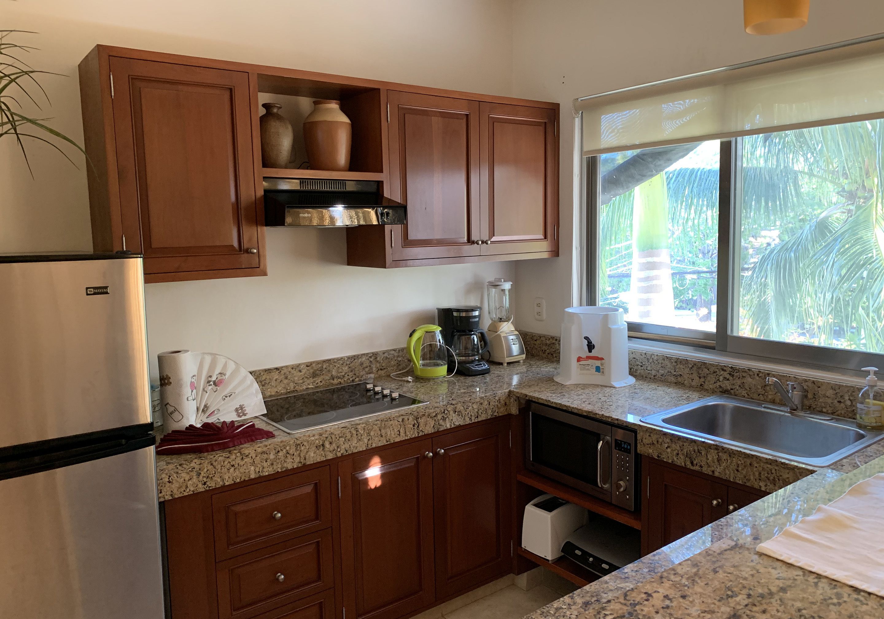A kitchen with wooden cabinets, a stainless steel refrigerator, a granite countertop, a sink, and a window with a view of greenery.