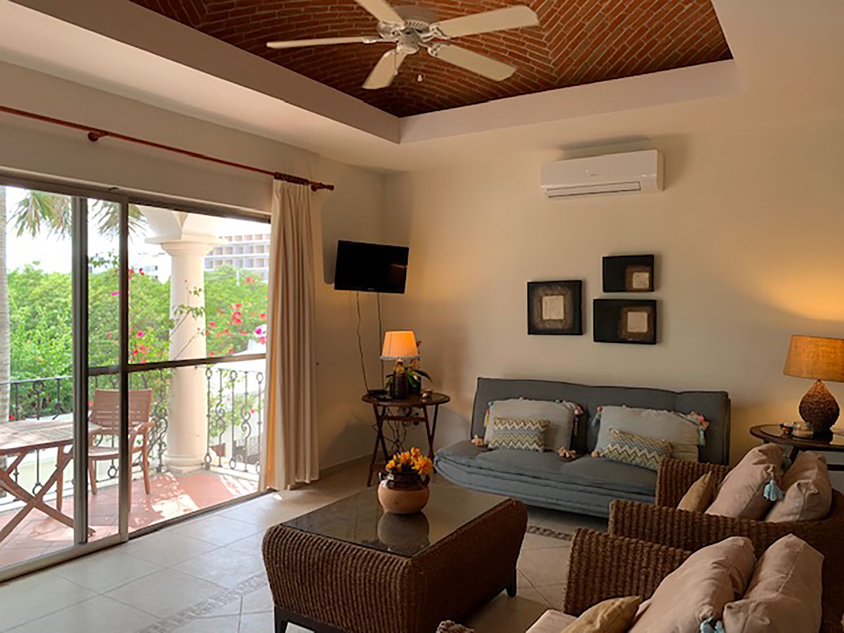 A spacious living room with a ceiling fan, a flat-screen TV mounted on the wall, a sofa with cushions, a coffee table, and a dining area with chairs.