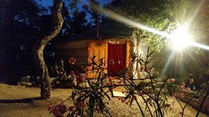A small, rustic wooden structure with a red door, surrounded by plants and rocks, illuminated by a single light source. - Au Soleil Mongol (Sorede)