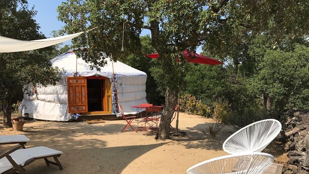 A yurt with a wooden door and a red umbrella, surrounded by trees and outdoor furniture including a white chair and a wooden table. - Au Soleil Mongol (Sorede)