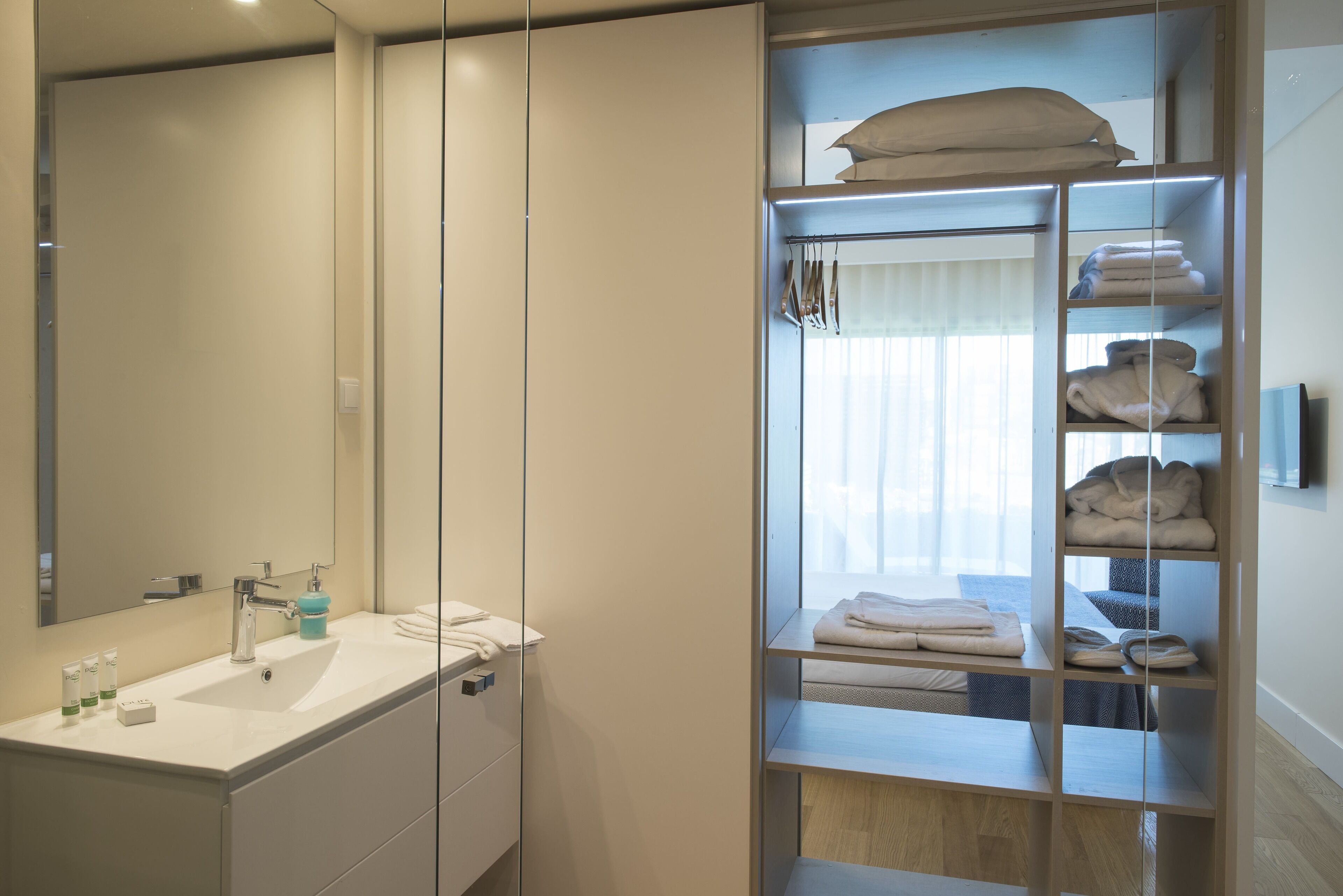 Ein modernes Badezimmer mit großem Spiegel, weißem Waschbecken und einem einbaubaren Kleiderschrank mit Glastüren und Ablagen für Handtücher und Kleidung.