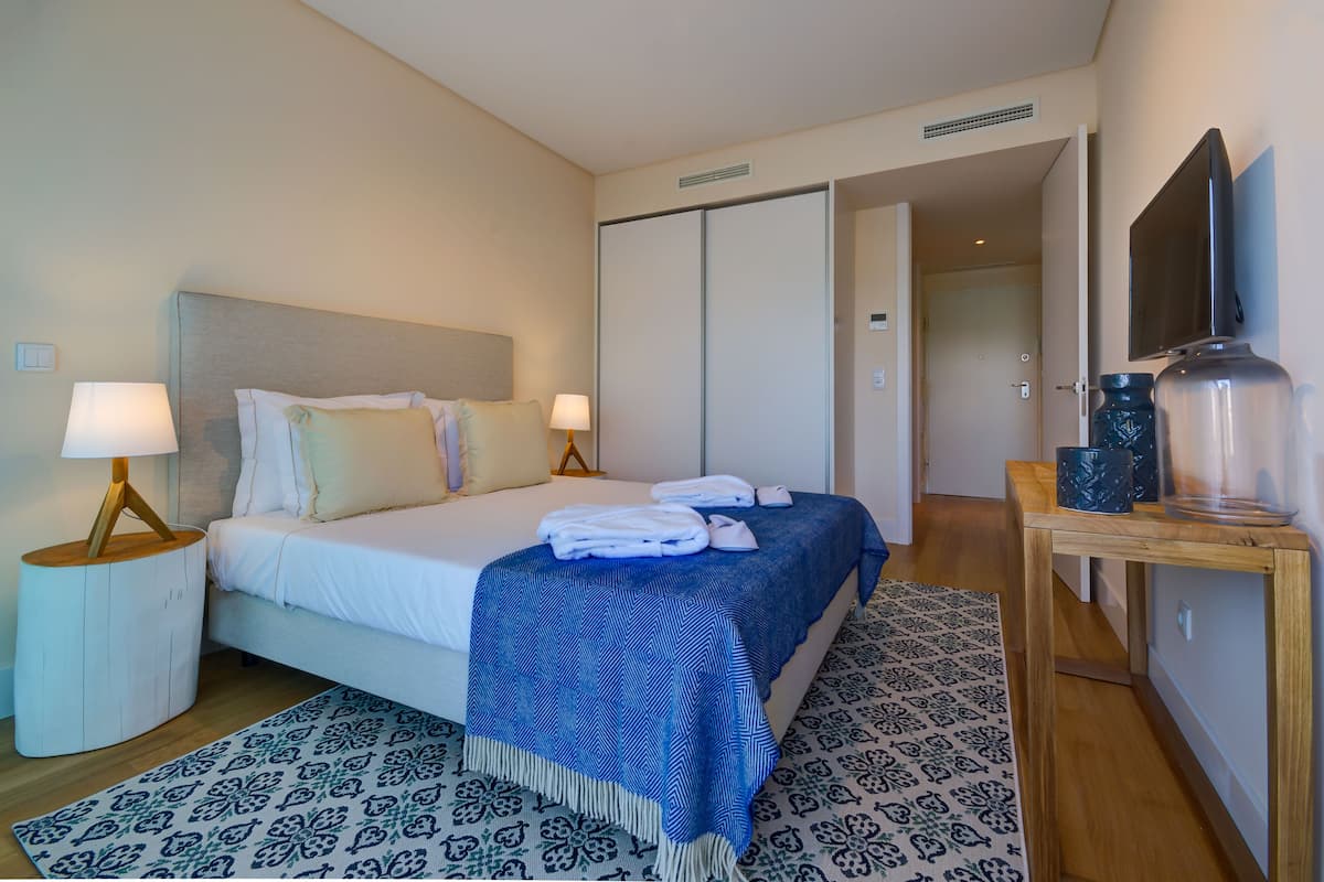 Una habitación de hotel con una cama, dos lámparas de noche, un tapete azul estampado, una mesita de madera con un jarrón y un televisor sobre un mueble.