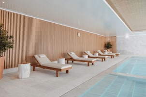 Uma área interna de piscina moderna, com paredes de painel de madeira, várias espreguiçadeiras e uma piscina azul cristalina.