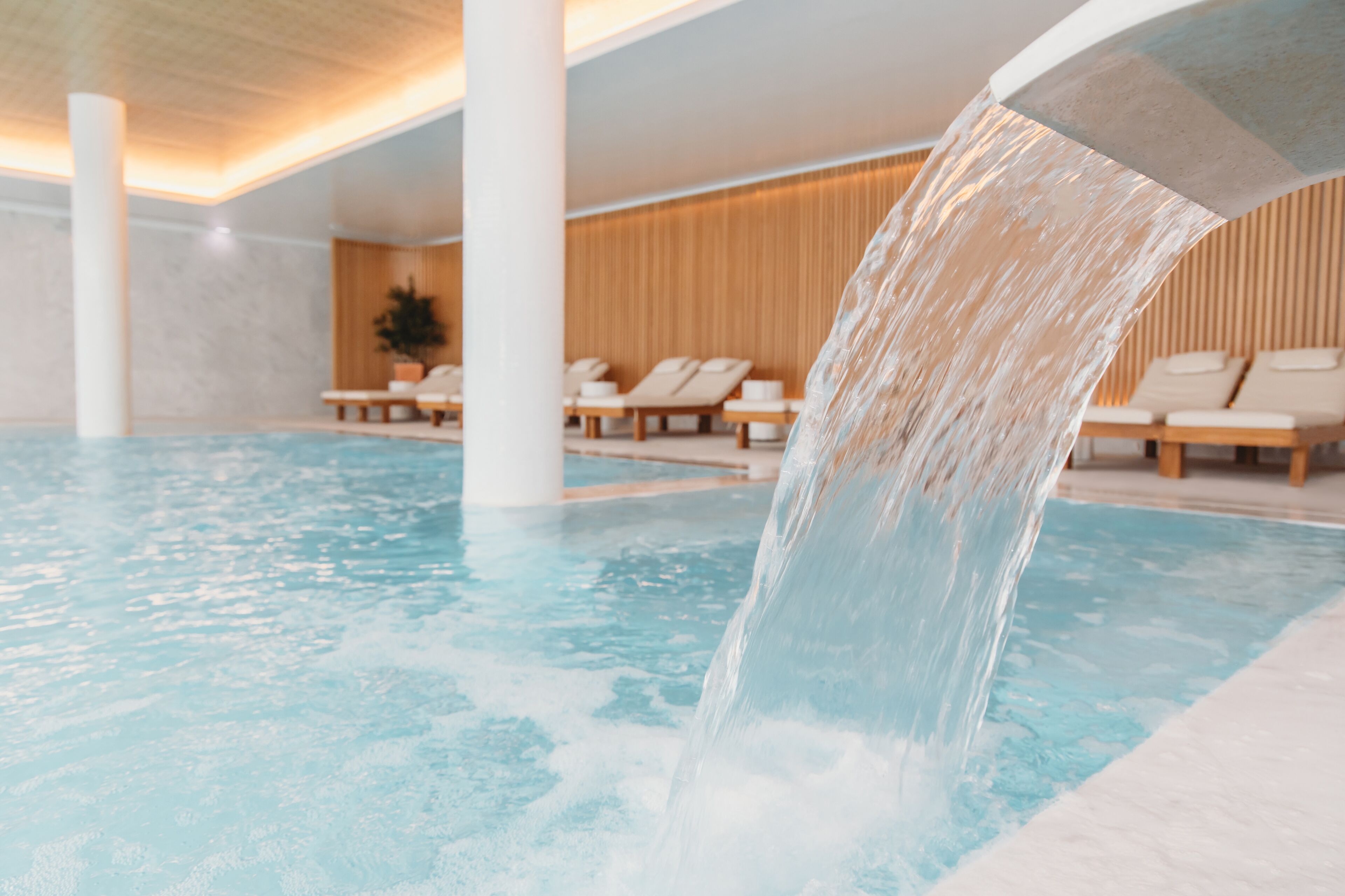 A pool with a water feature, lounge chairs, and a wooden wall in the background.