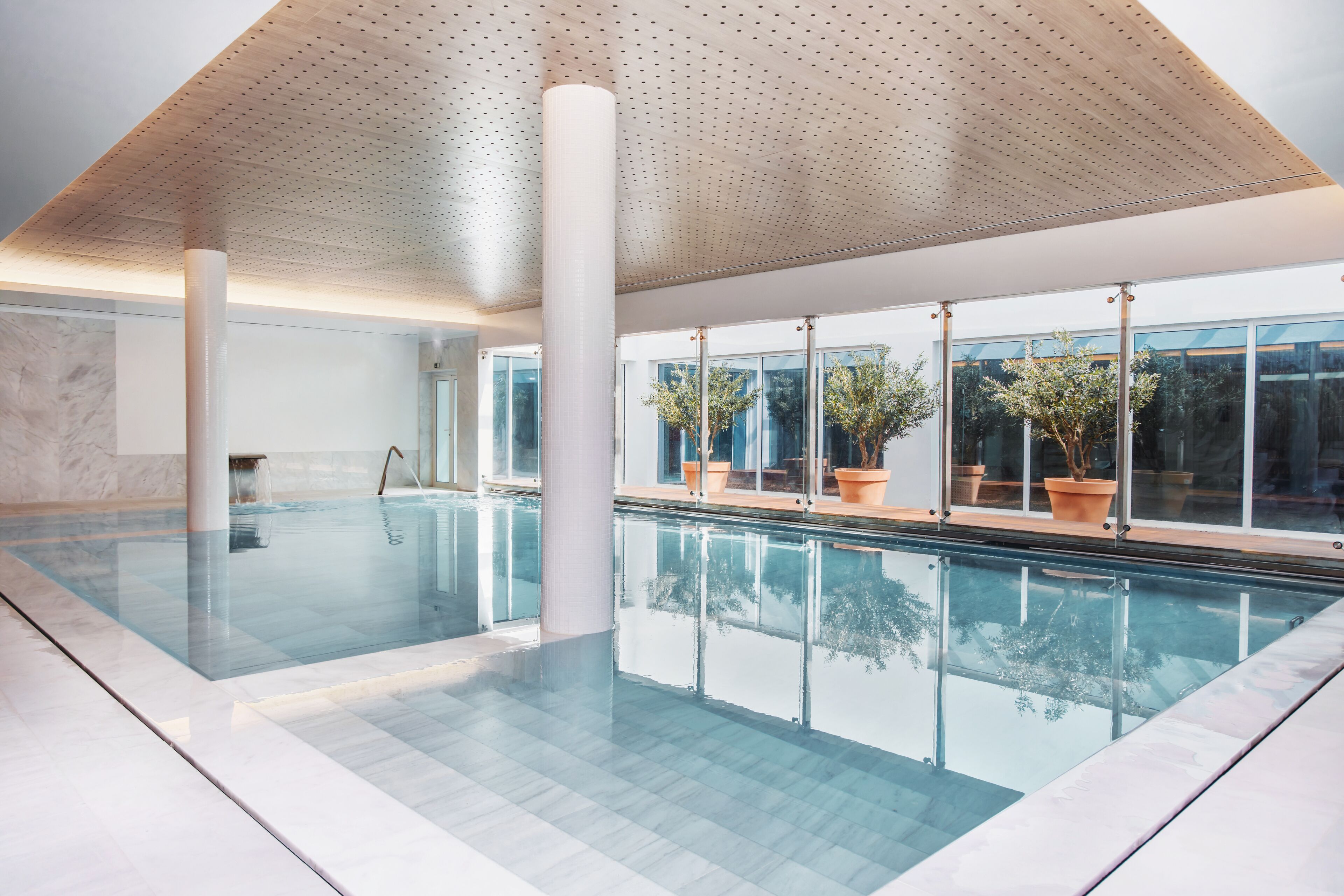 Una piscina interior moderna con paredes de vidrio transparente, grandes columnas y plantas en macetas.