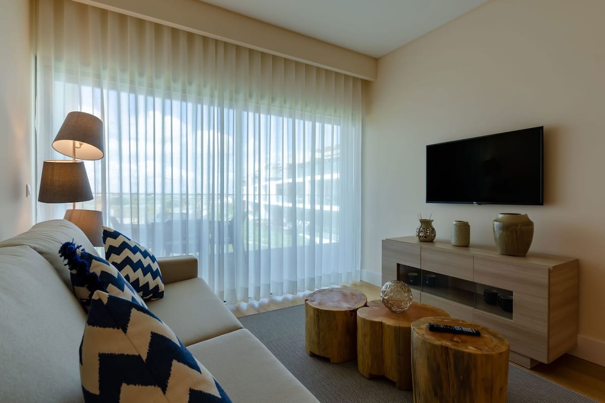 Una sala de estar moderna con sofá, televisor, consola de madera y un amplio ventanal con cortinas translúcidas.