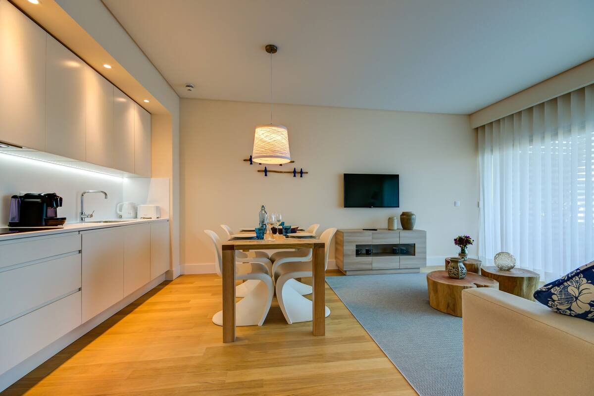Una cocina moderna con armarios blancos, una mesa de comedor con sillas, una lámpara colgante, un televisor de pantalla plana en la pared y un sofá con un cojín decorativo.