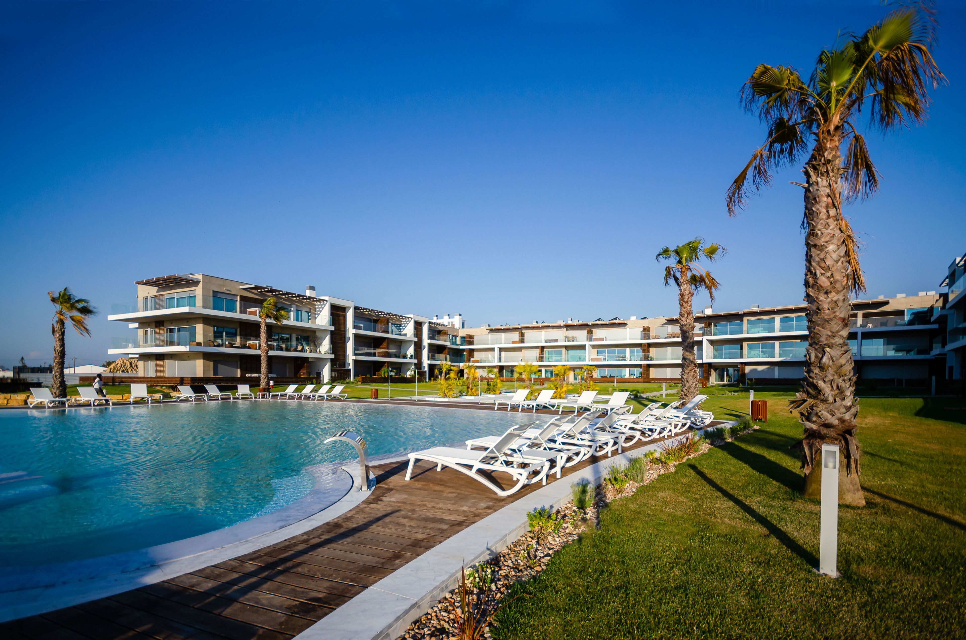 A modern hotel with a swimming pool, lounge chairs, and palm trees.
