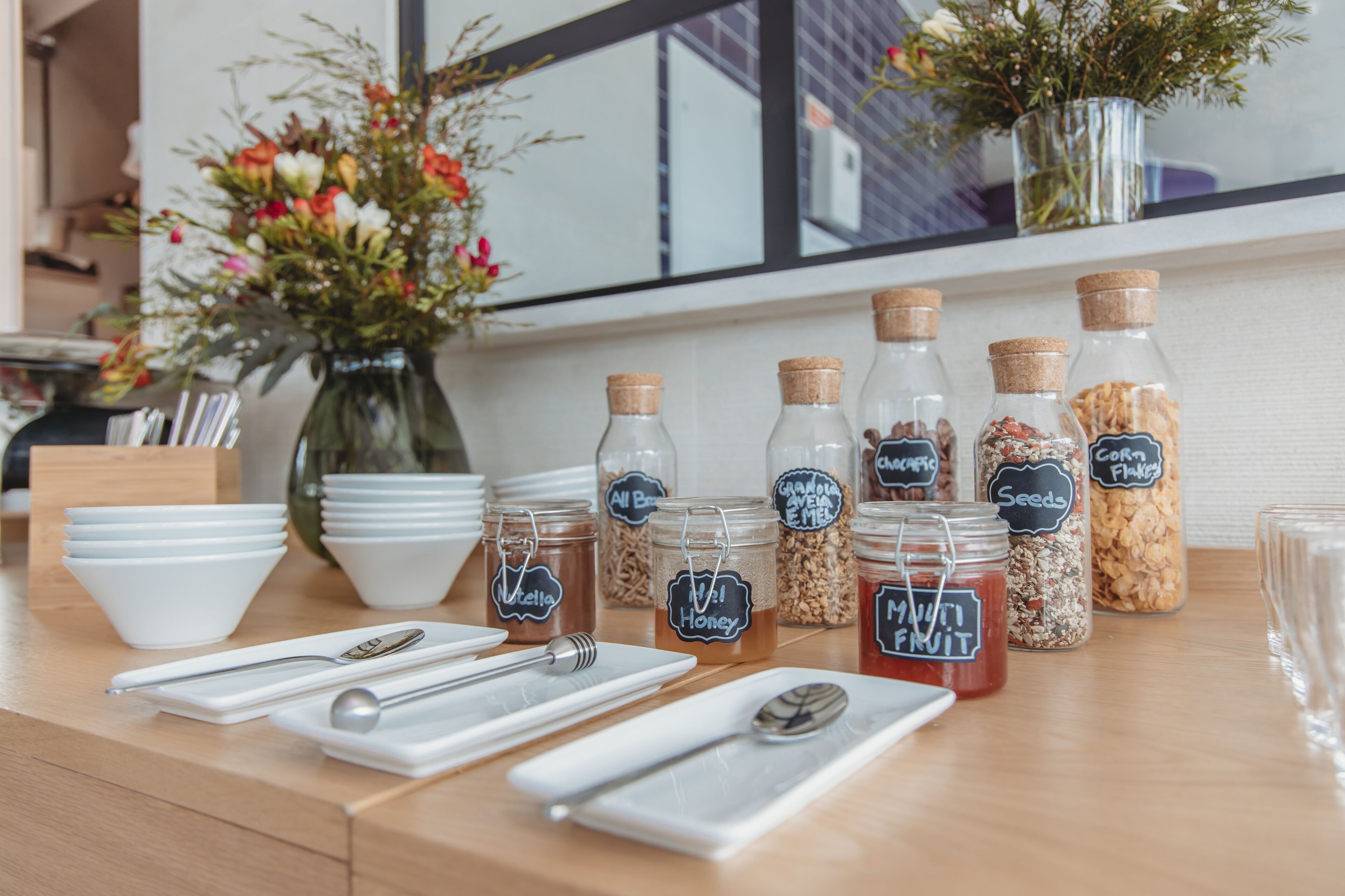 A breakfast setup with jars of honey, granola, and corn flakes, white bowls, and silverware on a wooden table.