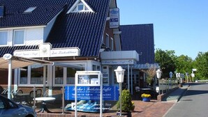 A building with a sloped roof, a covered outdoor seating area, and a sign advertising a restaurant. - Hotel zur Fischerklause (Butjadingen)