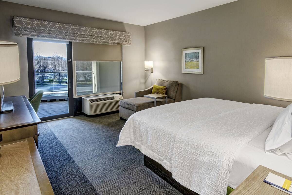 A hotel room with a large bed, a desk, a chair, a small table, a lamp, and a window with a view of trees.