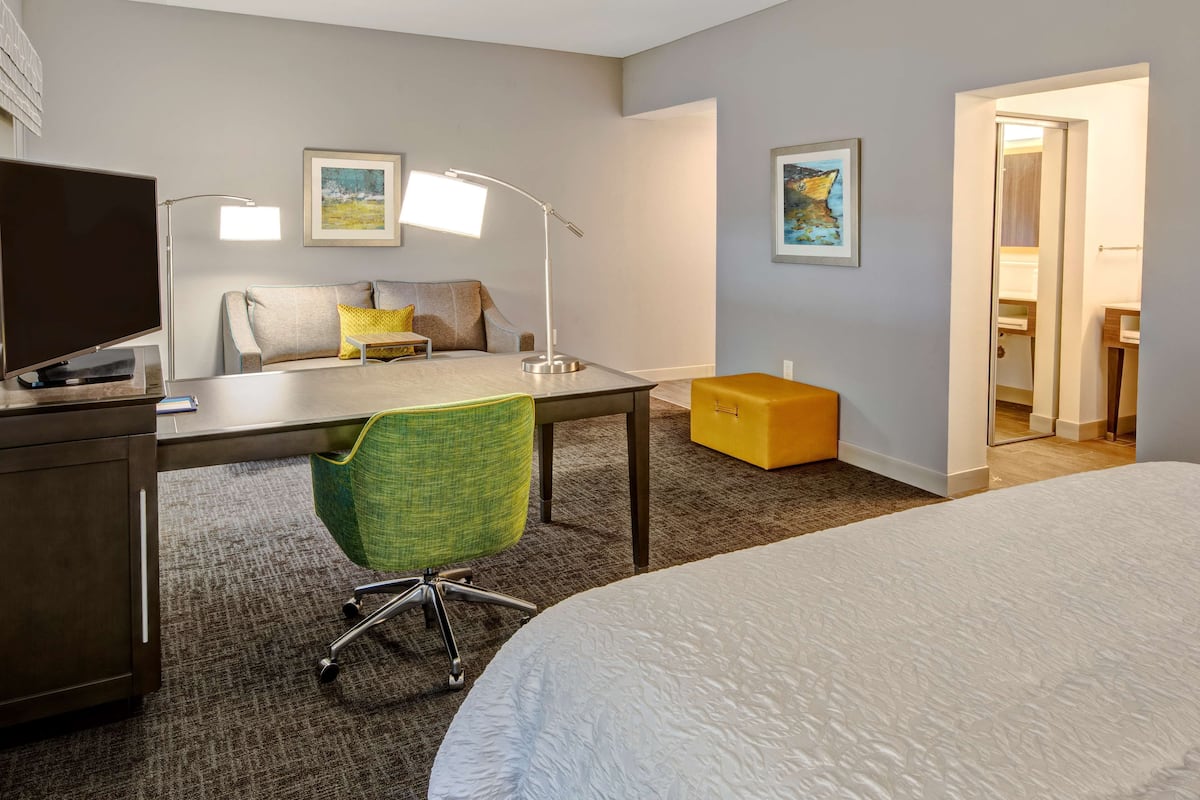 A hotel room with a bed, desk, chair, and a television. There are two paintings on the walls and a bathroom visible through an open door.