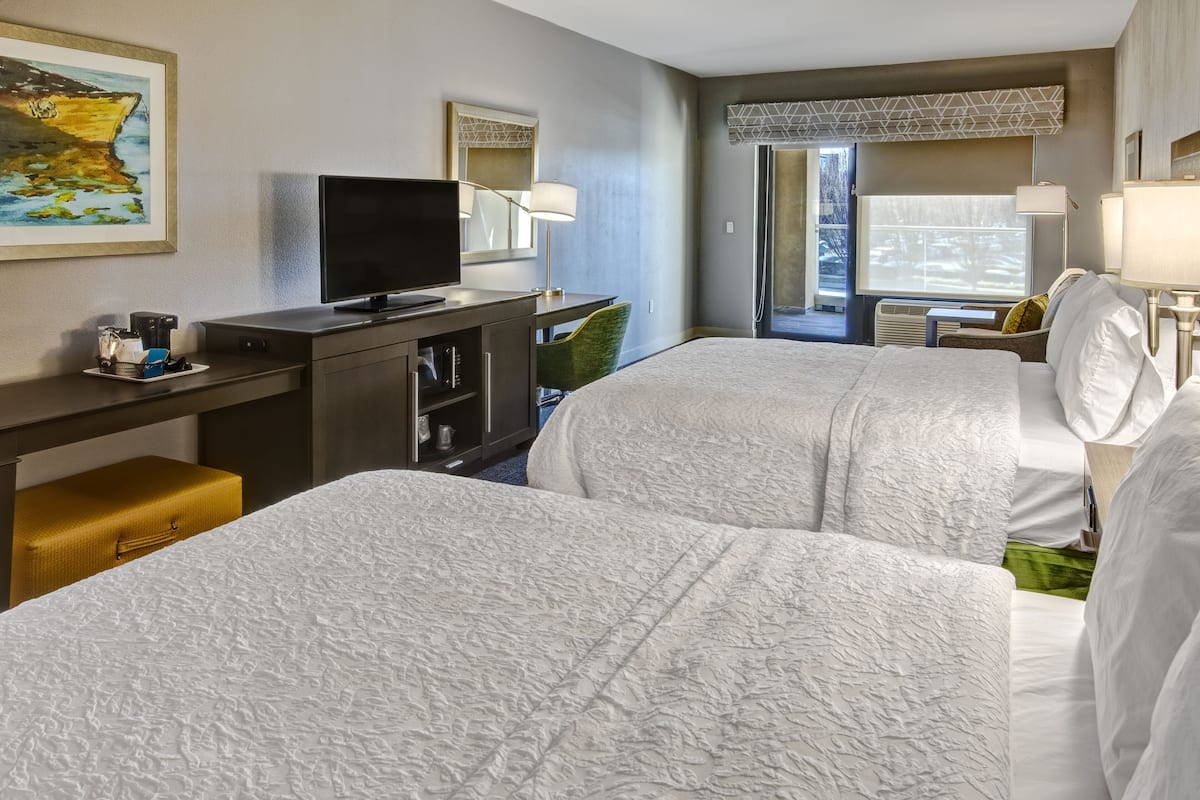 A hotel room with two beds, a desk with a television, and a window with a view of the outdoors.