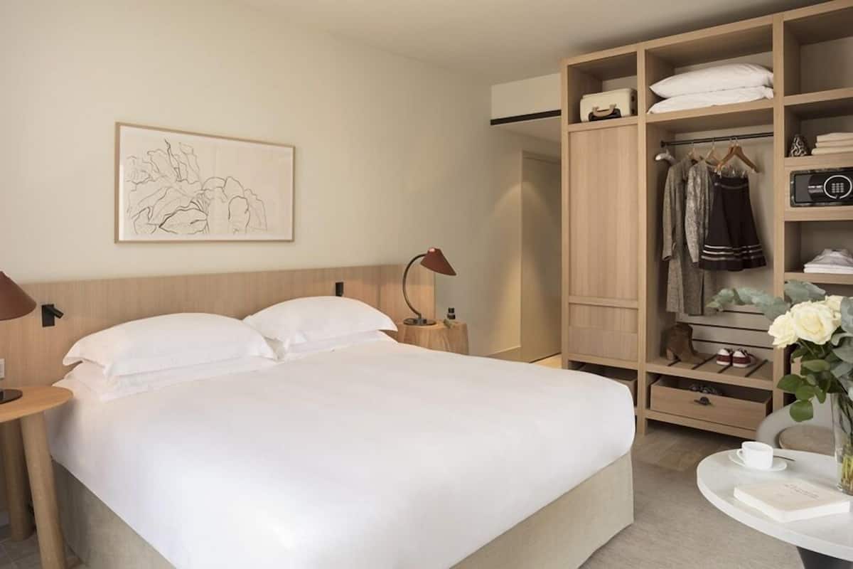 A hotel room with a large bed, a bedside table with a lamp, a wardrobe with hanging clothes, and a small table with a cup and book.