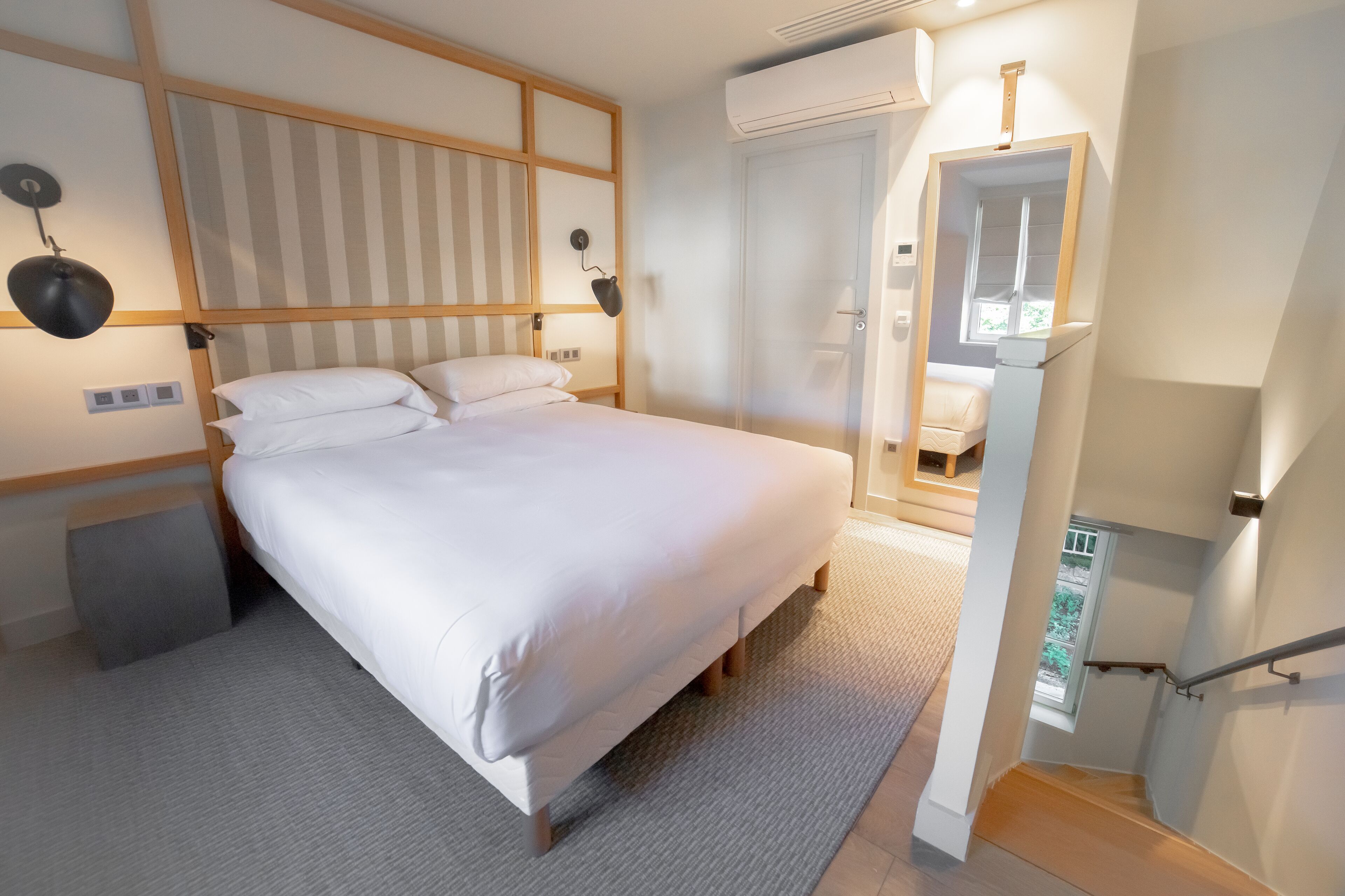 A bedroom with a bed, bedside lamps, a wooden headboard, a small bedside table, and a window.