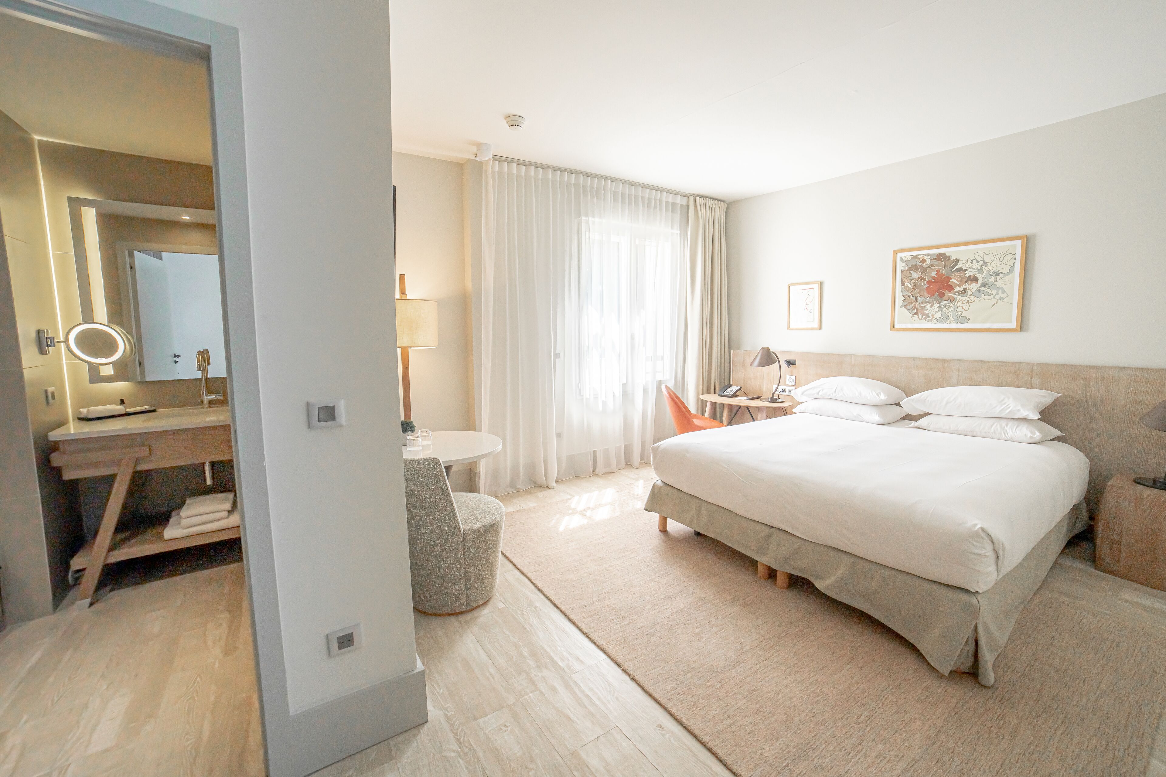 A hotel room with a bed, a desk with a mirror, a chair, a small table, and a window with curtains.