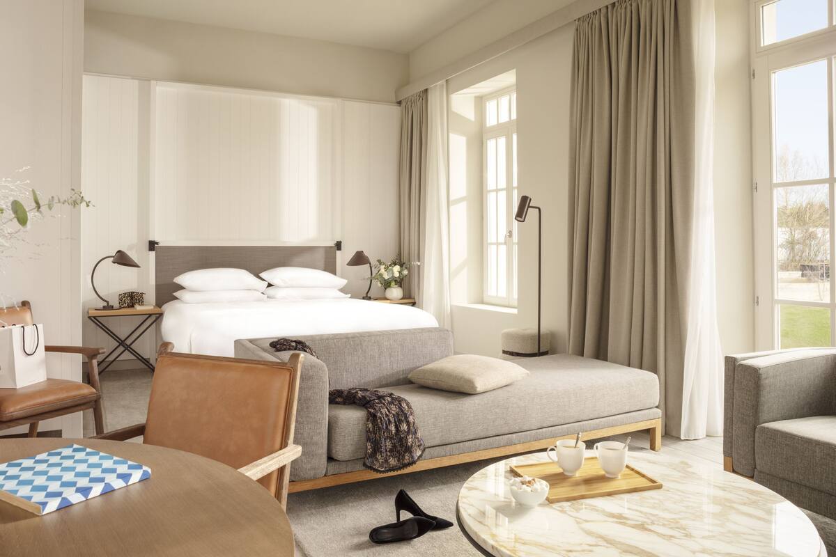 A hotel room with a bed, a sofa, a dining table, and a chair. There is a window with curtains, a plant, and a lamp on the bedside table.