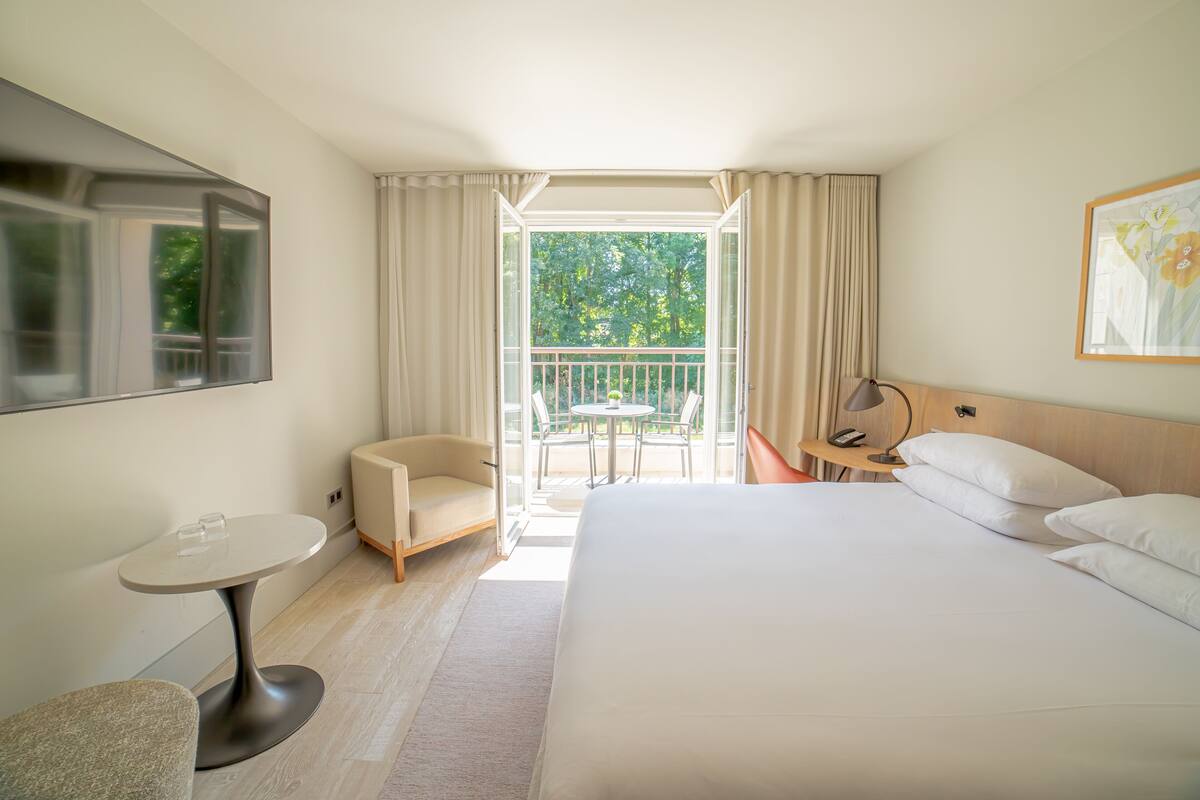 A hotel room with a bed, a small table, a chair, a desk, a TV, and a balcony with a view of trees.