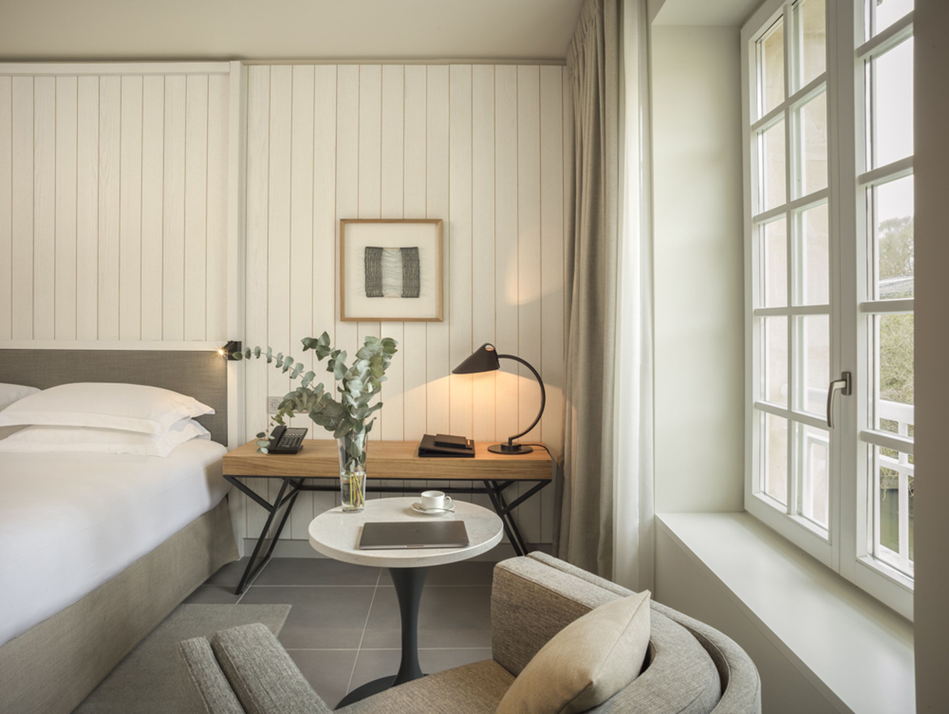 A hotel room with a bed, a small round table with a lamp, a chair, and a window with curtains.