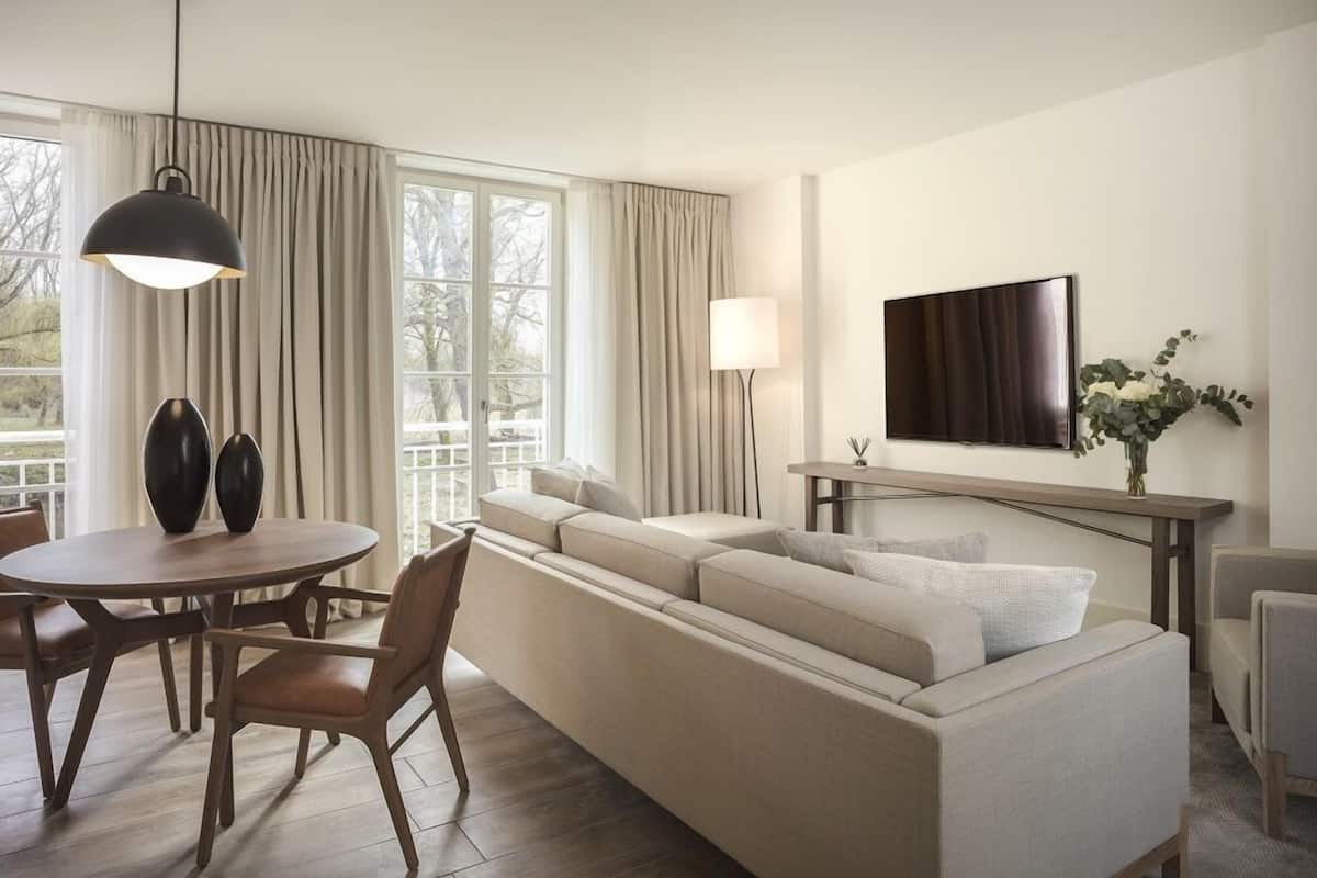 A modern hotel room with a large sofa, a dining table with chairs, a TV mounted on the wall, and a large window with curtains.