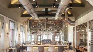 Ein großzügiges Restaurant mit hohen Decken, freiliegenden Lüftungsrohren und einer zentralen Bar. Die Tische sind ordentlich mit Stühlen bestückt, und der Boden ist aus Holz.
