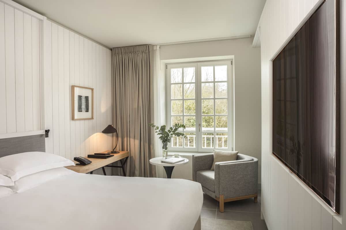 A hotel room with a bed, a desk with a lamp, a chair, a small table with a plant, and a large window with a view of trees.
