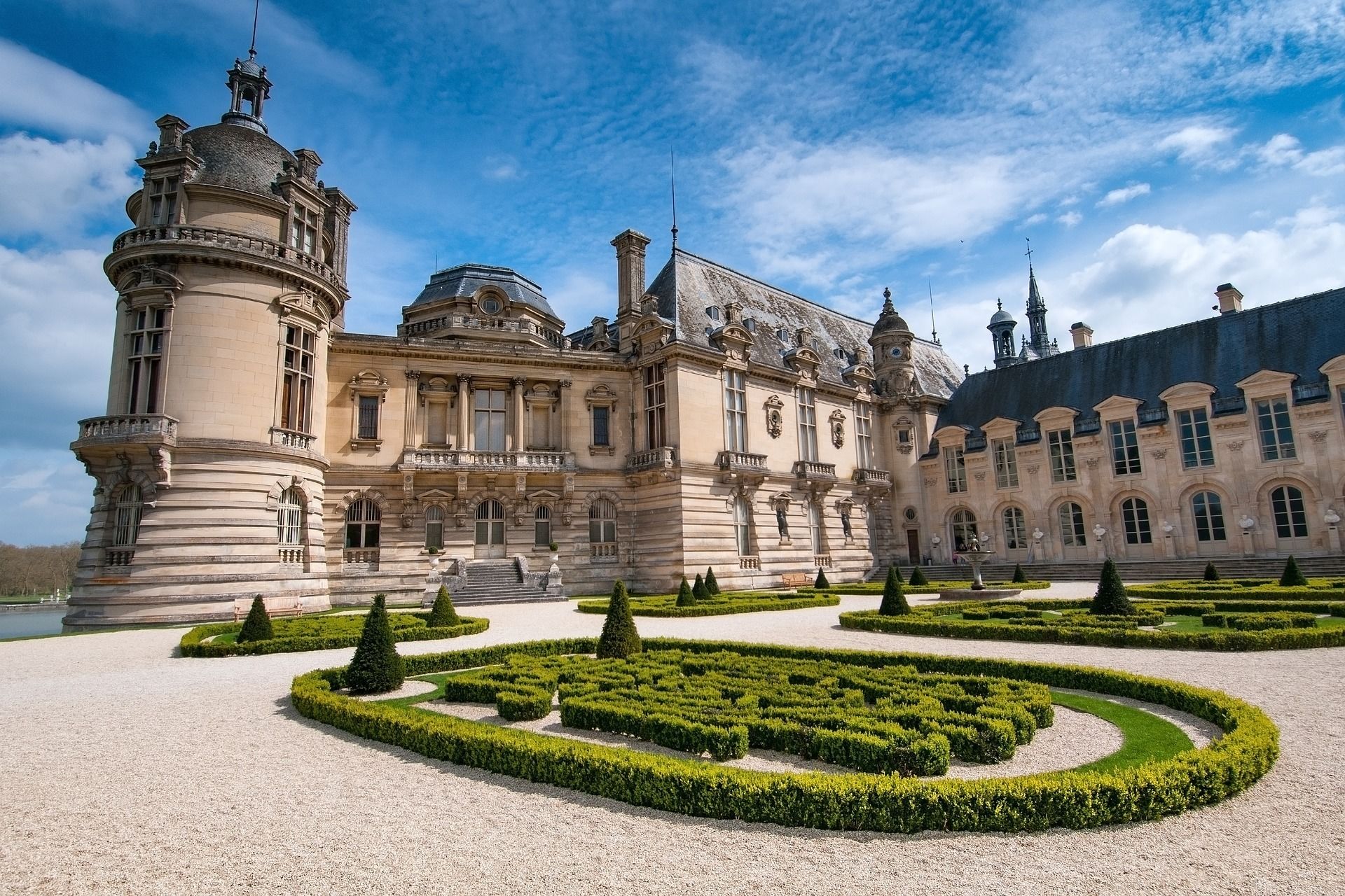 A grand historic building with symmetrical architecture, featuring a central courtyard with geometric hedges and a fountain.