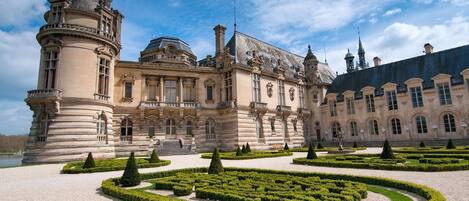 A grand historic building with symmetrical architecture, featuring a central courtyard with geometric hedges and a fountain.