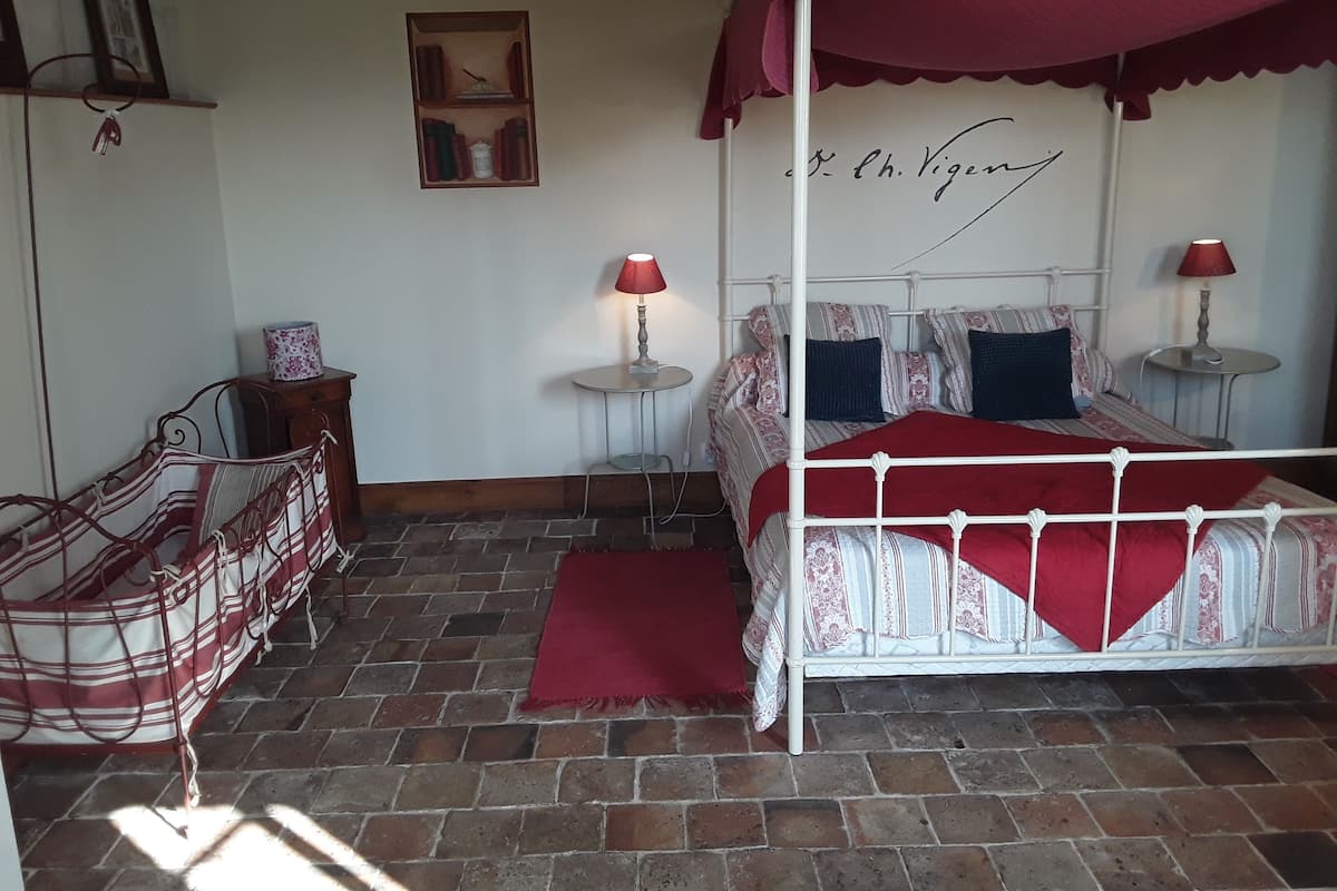 Un lit à baldaquin à quatre poteaux surmonté d’une couverture rouge, deux tables de chevet avec des lampes, une petite armoire en bois et un sol décoré d’un motif imitant des briques.