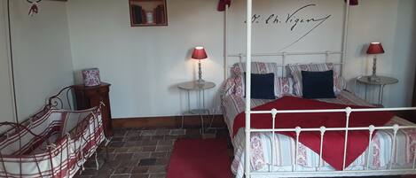 Una cama con dosel y cuatro columnas, con un edredón rojo, dos mesitas de noche con lámparas, un pequeño armario de madera y un suelo con diseño de ladrillo.