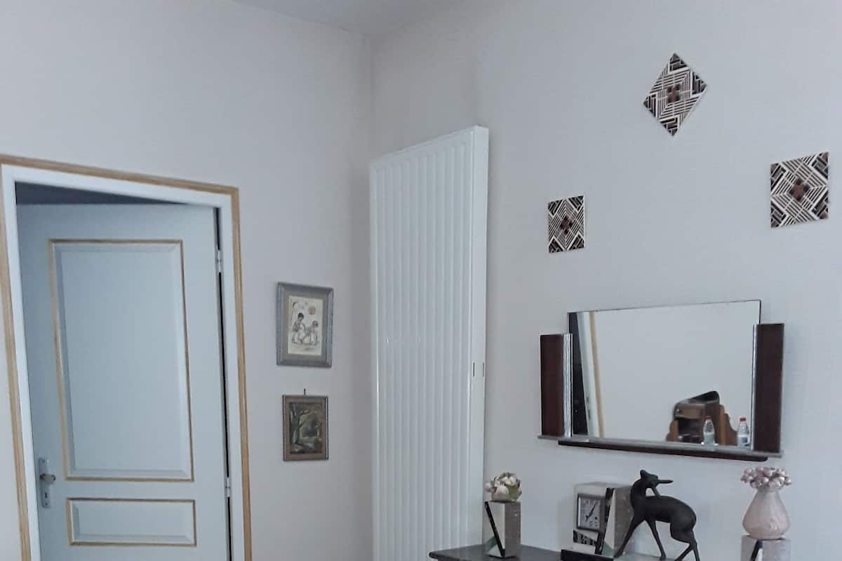 Une pièce avec un plancher en bois, une porte blanche, une chaise rouge, une chaise noire, un radiateur blanc, une cheminée en marbre, un miroir et des carreaux décoratifs sur les murs.