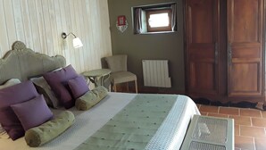 Een slaapkamer met een bed, kussens, een klein tafeltje, een stoel, een radiator en een houten garderobekast.