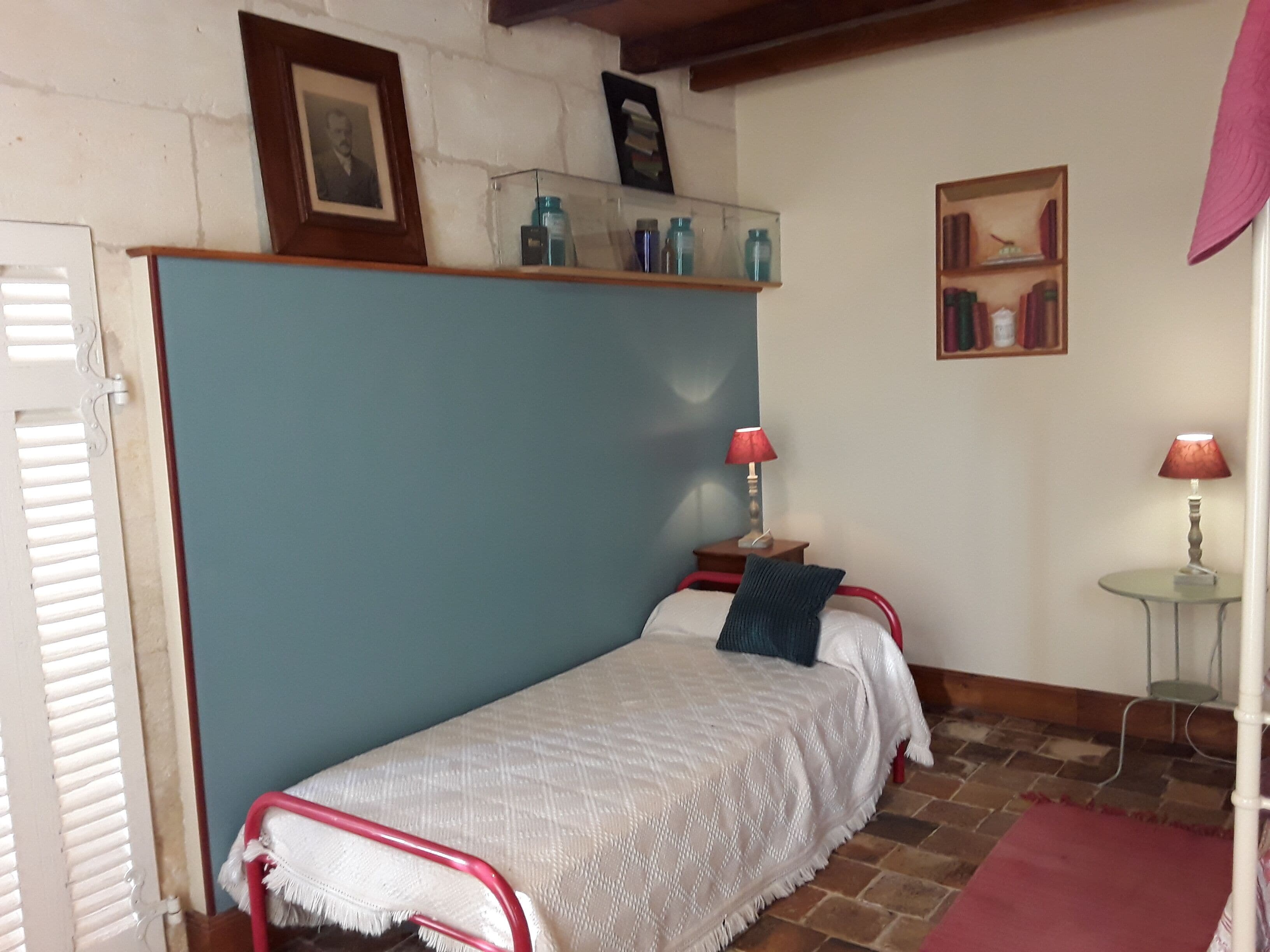 Une petite chambre à coucher, chaleureuse et accueillante, avec un lit simple, une table de chevet, une lampe, une étagère avec des bocaux et un tableau encadré au mur.