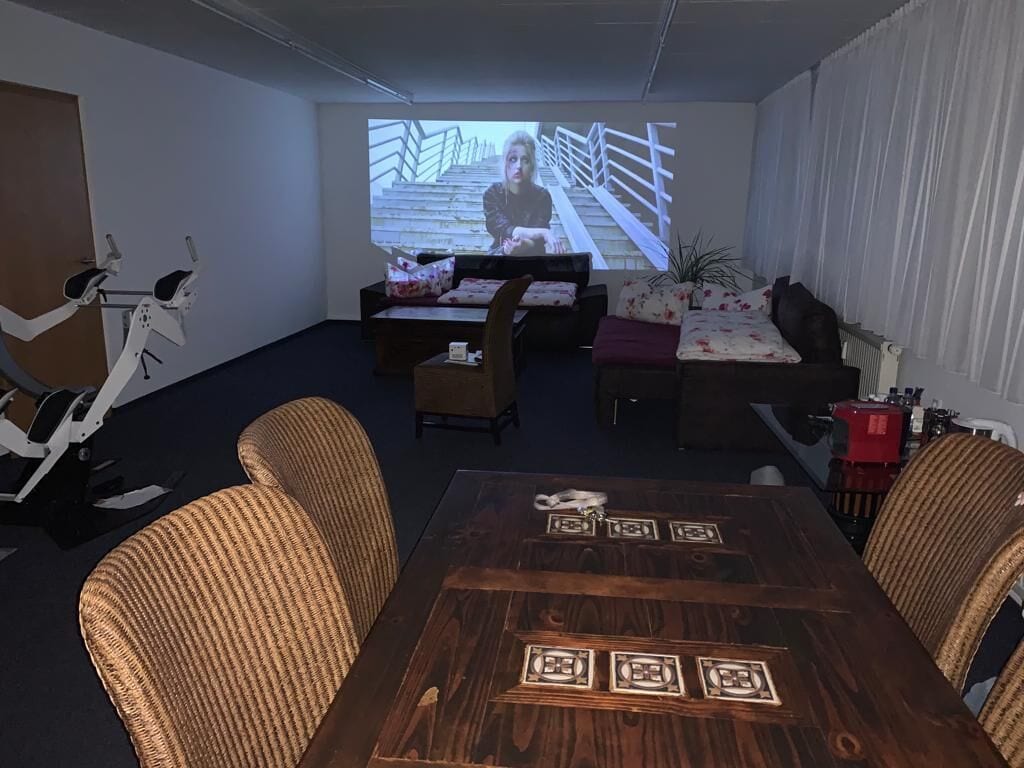 Una habitación con una mesa de comedor de madera, sillas de mimbre, un sofá y una pantalla de proyección mostrando una escena.