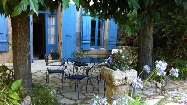 A stone patio with a table and chairs, surrounded by trees and flowering plants. - Aux Pots Bleus (Nissan-lez-Enserune)