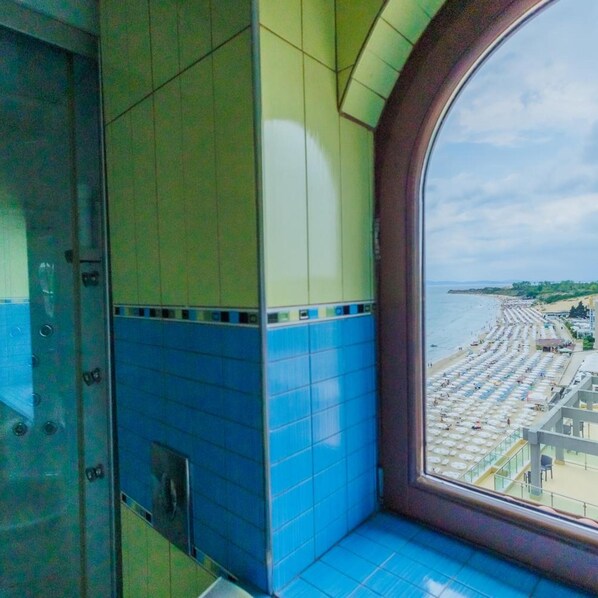 A room with a large window overlooking a beach with umbrellas and people. - Mirage Hotel (Nessebar)