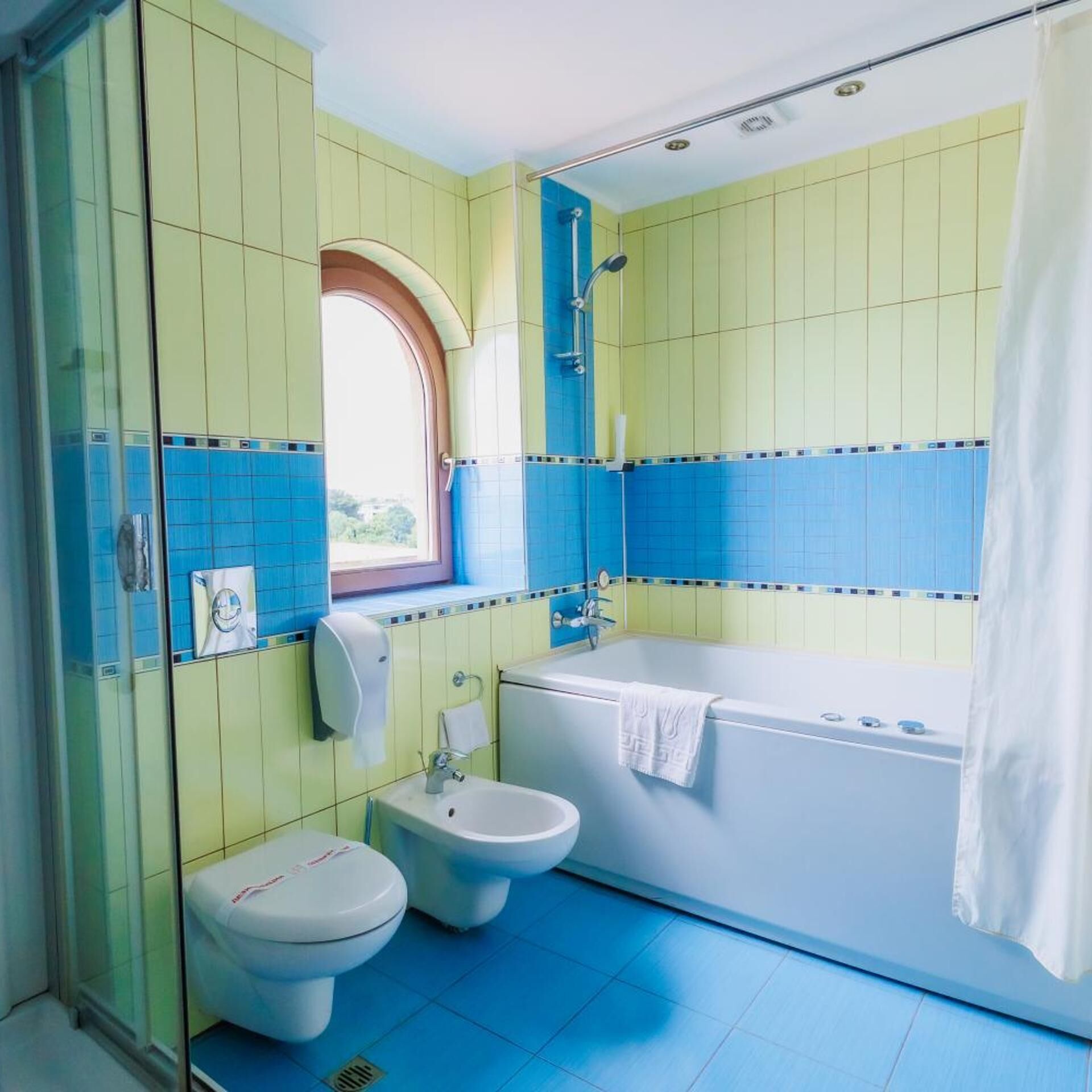 Un baño con bañera, dos inodoros, un secador de manos y una ventana.