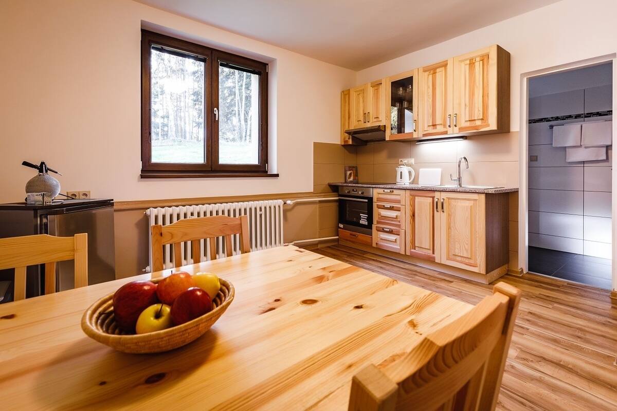 Una cocina con armarios de madera, una mesa de comedor con un frutero y una ventana con vistas a árboles.