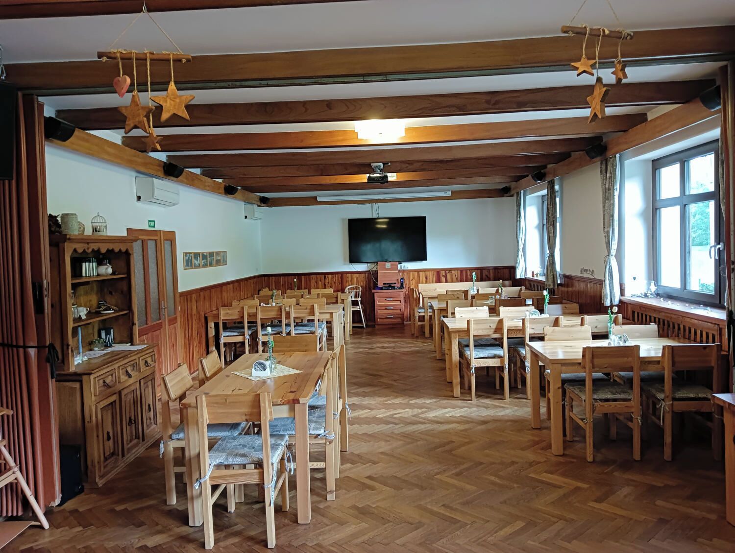 A large hall with wooden tables and benches, a television screen, and a herringbone floor pattern.