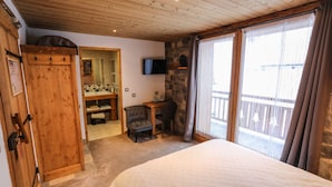 Un dormitorio con una cama grande, techo de madera, un escritorio con silla, un televisor y un balcón con cortinas.