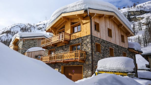 Un chalet de madera con paredes de piedra, balcones y un techo cubierto de nieve, en un paisaje montañoso.