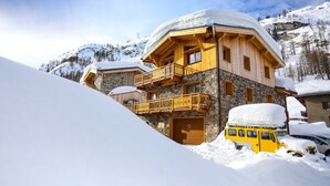 Una cabaña de madera con base de piedra, un balcón y un techo cubierto de nieve, rodeada de nieve y montañas.