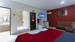 Habitación de hotel con cama, televisión, sofá y baño con ducha, y una pared roja.