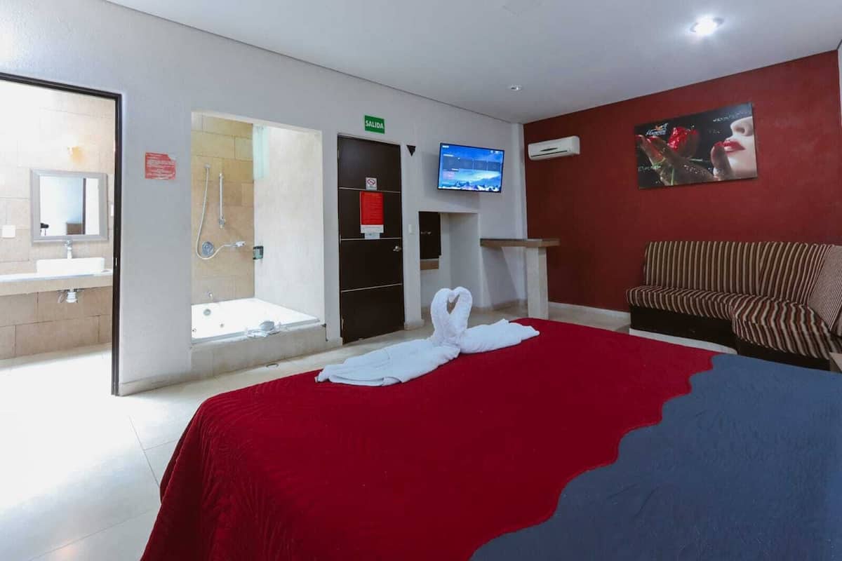 A hotel room with a bed, a TV, a sofa, a bathroom with a shower, and a red wall.