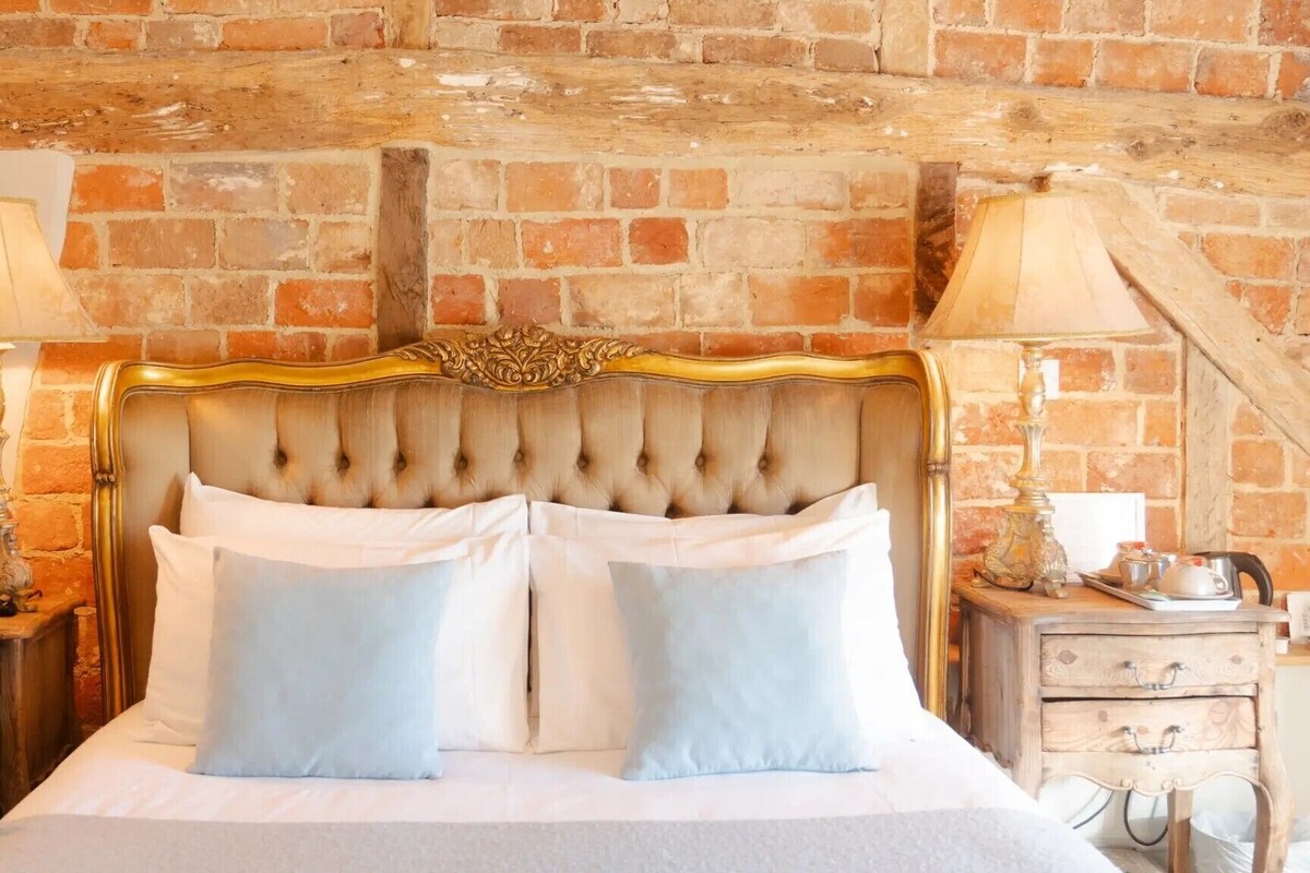 A bed with a tufted headboard, two bedside tables with lamps, a small wooden nightstand, and a brick wall.