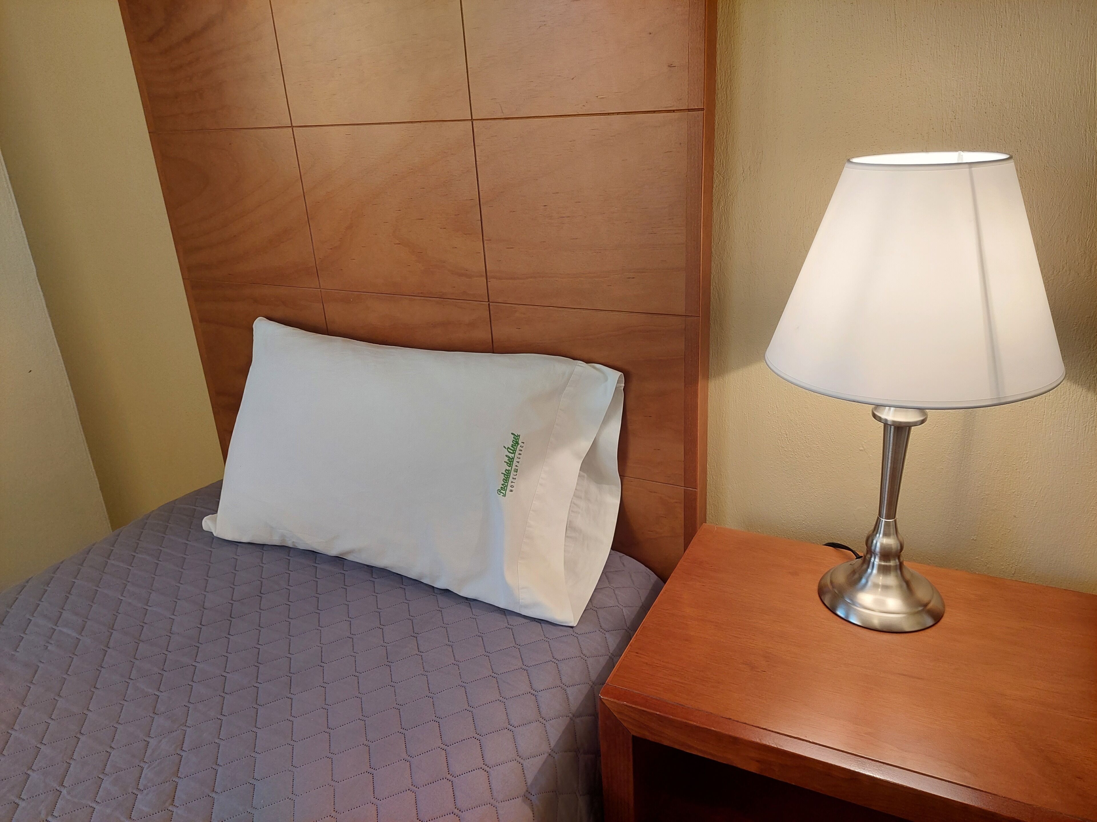 Una cama bien hecha con una colcha estampada, un almohadón blanco con texto verde y una mesita de noche con una lámpara.