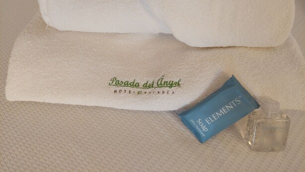 Una toalla blanca doblada con cuidado, un jabón azul de la marca Elements y una botella transparente de champú sobre una superficie texturizada.
