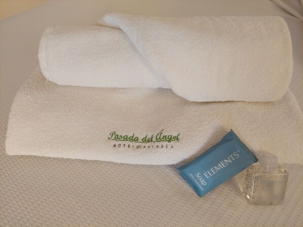 Una toalla blanca doblada con cuidado, un jabón azul de la marca Elements y una botella transparente de champú sobre una superficie texturizada.