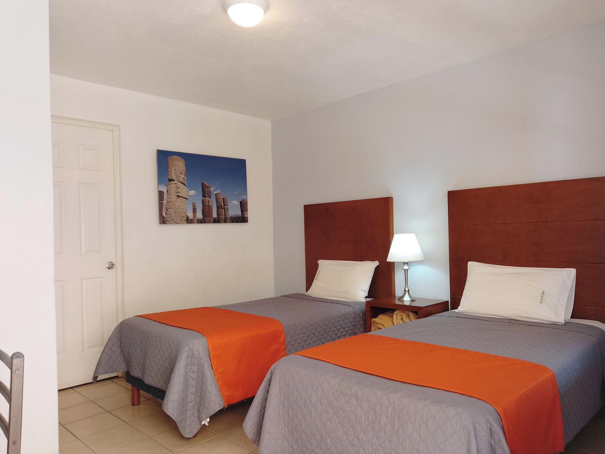 Habitación de hotel con dos camas, una mesita de noche con lámpara, un cuadro en la pared y una puerta.